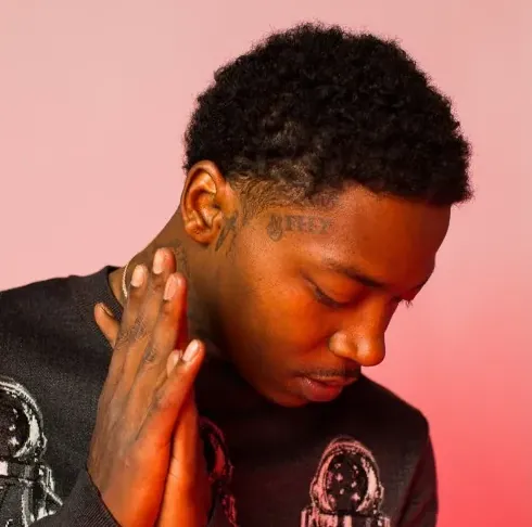 2FeetBino in a black graphic sweater, eyes lowered, hands pressed together against a pink backdrop