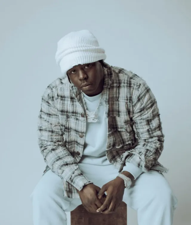 BabyDrill seated in a white beanie and plaid jacket against a pale grey backdrop